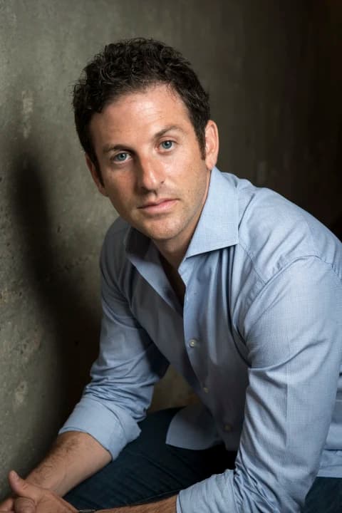 Man with dark curly hair and blue eyes wearing a light blue button-down shirt.